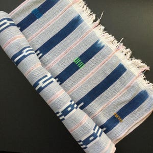 May include: Rolled and unfolded textile with a striped pattern. The fabric features alternating bands of blue, white, and pink, with fringed edges. The design includes small, colorful accents in green and yellow. The textile is likely used for home decor.
