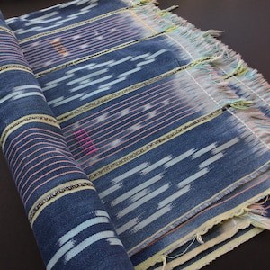 May include: Rolled blue fabric with a striped pattern. The fabric features horizontal stripes in pink and yellow, with light blue and white brushstroke-like designs. The edges are fringed with a mix of colors. The fabric is partially rolled up.