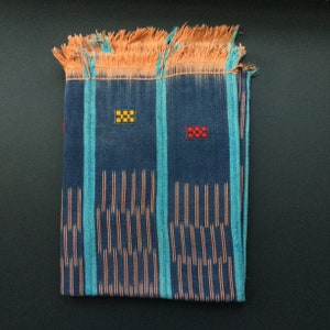 May include: Folded blue textile with turquoise stripes and orange fringe. The fabric features vertical orange lines and small embroidered squares in yellow and red. A decorative piece.