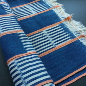 May include: A rolled-up, indigo blue textile with white horizontal stripes and orange accent stripes. The fabric has a fringed edge and a textured appearance, suggesting a woven or hand-crafted item. The design is simple and geometric.