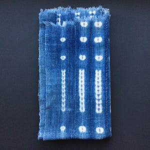 Könnte beinhalten: Indigoblauer Stoff mit weißen Punkt- und Linienmustern. Der Stoff hat einen Fransenrand und ein strukturiertes Aussehen. Das Design zeigt Reihen weißer Punkte und vertikaler Linien, wodurch ein geometrisches Muster entsteht. Der Stoff ist gefaltet und zeigt das Design.