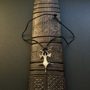 May include: A silver pendant necklace with a geometric design, suspended on a black cord. The pendant is displayed against a carved wooden background with geometric patterns. The necklace is a piece of jewelry.