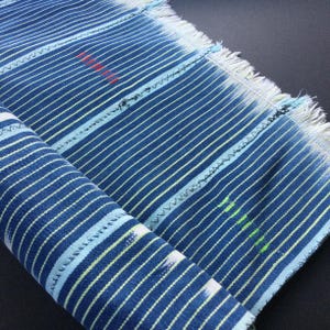 May include: Close-up of a rolled-up blue and white striped fabric with a fringed edge. The fabric has thin white and light blue stripes on a dark blue background. There are small red and green markings on the fabric.