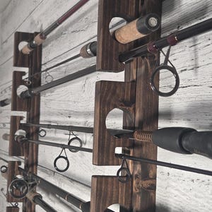 Può includere: Un supporto in legno tinto scuro per canne da pesca montato su una parete a pannelli di legno bianco. Il supporto contiene diverse canne da pesca di varie lunghezze e colori. Le canne sono fissate in aperture circolari.