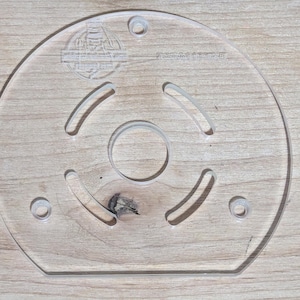 Op de afbeelding: Een heldere, cirkelvormige acryl freesbasisplaat met een centraal gat en drie gebogen sleuven. Het heeft drie kleinere gaten en een geëtst logo. De plaat is ontworpen voor houtbewerking.