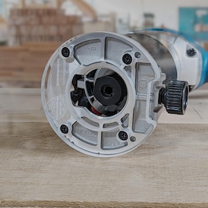 May include: A close-up of a silver router base with a black center and a clear plastic insert. The base has a black screw on the side and the text "391990 7227" printed on it.