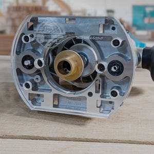 Puede incluir: Una plantilla de router de plástico transparente con un inserto de latón y una base de metal plateado. La plantilla tiene un logotipo en forma de estrella y el texto "MK18004-2P-F" impreso en ella.