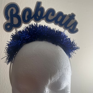 Puede incluir: Una diadema azul y blanca con la palabra "Bobcats" en letras azules de purpurina. La diadema está decorada con  escarcha azul.