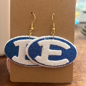 Puede incluir: Un par de pendientes ovalados de purpurina azul y blanca con ganchos dorados. Los pendientes presentan las letras "IE" en blanco.