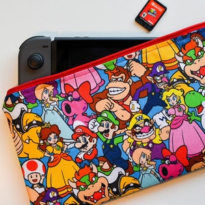 May include: A red zippered pouch with a blue background and a colourful pattern of various Nintendo characters, including Mario, Luigi, Donkey Kong, and Princess Peach. The pouch is made of fabric and has a zipper closure.