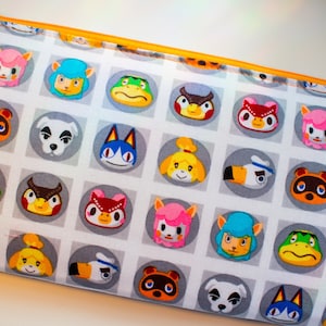 May include: A white zippered pouch with a repeating pattern of animal faces from the Animal Crossing video game. The faces are in circles on a gray background with white squares.