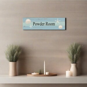 May include: A light blue wooden sign with white seashells and starfish accents. The sign reads "Powder Room".