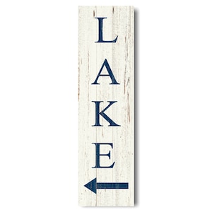 May include: A rectangular wooden sign with a distressed white background and navy blue lettering. The word "LAKE" is vertically stacked, with an arrow pointing to the left below the word. The sign has a rustic, weathered appearance.