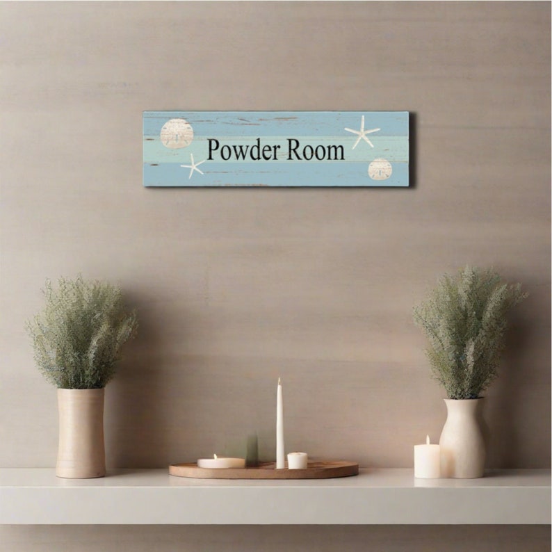 May include: A light blue wooden sign with white seashells and starfish accents. The sign reads "Powder Room".