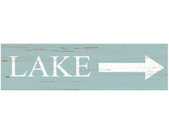 May include: A rectangular wooden sign with a distressed light blue finish featuring the word LAKE in large white capital letters followed by a white arrow pointing to the right. The surface shows horizontal wood grain and weathered texture.