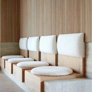 May include: A row of five white cushioned benches with wooden frames against a wall of vertical wooden slats.