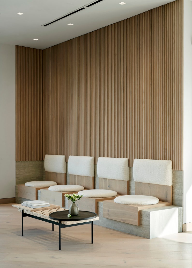 May include: A waiting area with a wooden slat wall and a row of white cushioned benches with wooden bases. A black metal coffee table with a wooden top sits in front of the benches.