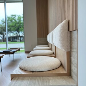 May include: A row of white cushioned seats with wooden frames against a wall with wood paneling. The seats are in a waiting area with a window in the background.