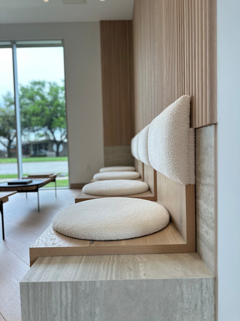 May include: A row of white cushioned seats with wooden frames against a wall with wood paneling. The seats are in a waiting area with a window in the background.