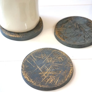 May include: A white ceramic mug rests on a gray coaster with gold accents. Two additional coasters are visible, each with a distressed, textured appearance. The coasters are round and appear to be made of a solid material.