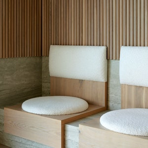May include: Two white cushioned seats with wooden frames are built into a wall with vertical wooden slats. The seats are set on a light gray stone surface.