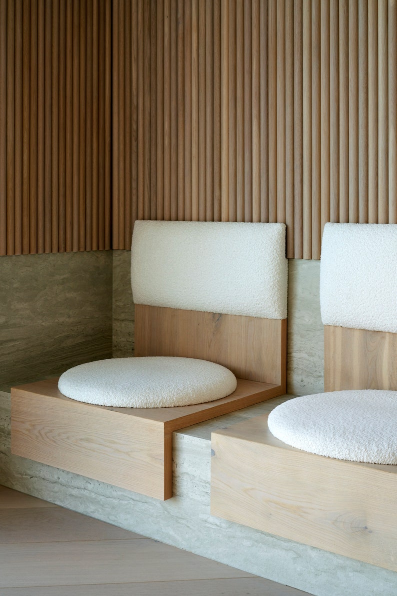 May include: Two white cushioned seats with wooden frames are built into a wall with vertical wooden slats. The seats are set on a light gray stone surface.
