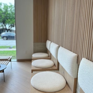 May include: A row of four white cushioned chairs with wooden frames and backs. The chairs are arranged in a waiting area with a wood slat wall behind them.