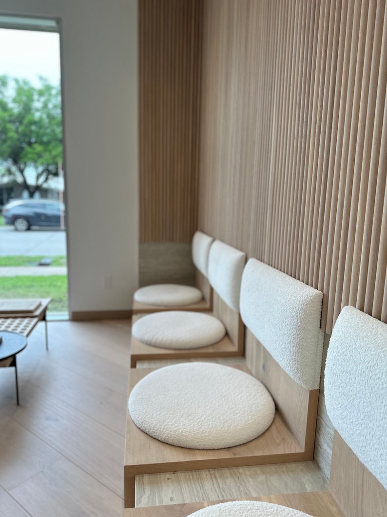 May include: A row of four white cushioned chairs with wooden frames and backs. The chairs are arranged in a waiting area with a wood slat wall behind them.