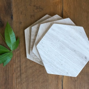 May include: A set of four hexagonal coasters made of light beige stone with a natural, linear grain pattern. The coasters are stacked in a slightly offset arrangement, showcasing the smooth, flat surfaces and the distinct six-sided geometric shape.