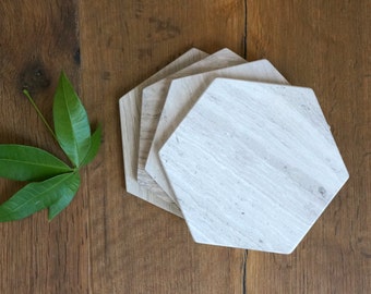 May include: A set of four hexagonal coasters made of light beige stone with a natural, linear grain pattern. The coasters are stacked in a slightly offset arrangement, showcasing the smooth, flat surfaces and the distinct six-sided geometric shape.