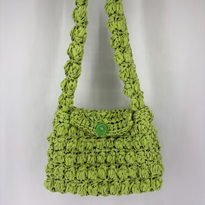 Può includere: Una borsa verde lime con un design strutturato e intrecciato. La borsa presenta una chiusura con patta assicurata da un bottone verde abbinato. La tracolla è anch'essa realizzata con lo stesso materiale intrecciato.