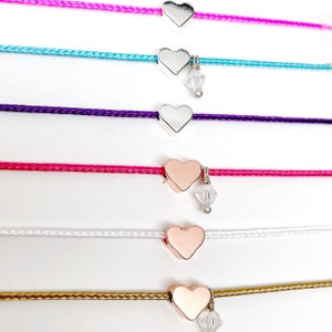 May include: Six heart charm bracelets with different colored cords. The cords are pink, turquoise, purple, hot pink, white, and gold. The heart charms are silver and rose gold. Some of the charms have a small crystal hanging from them.