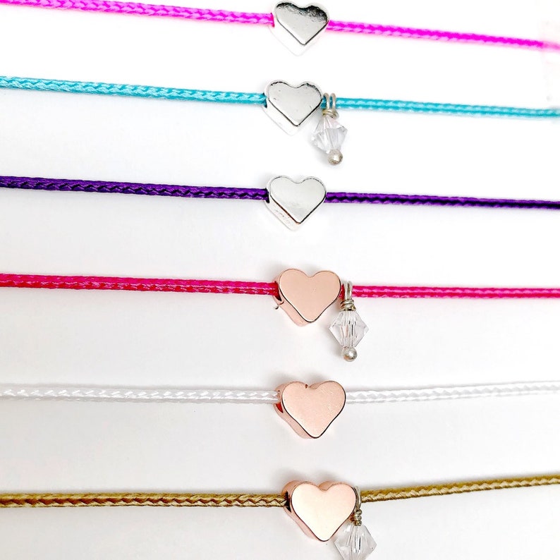 May include: Six heart charm bracelets with different colored cords. The cords are pink, turquoise, purple, hot pink, white, and gold. The heart charms are silver and rose gold. Some of the charms have a small crystal hanging from them.