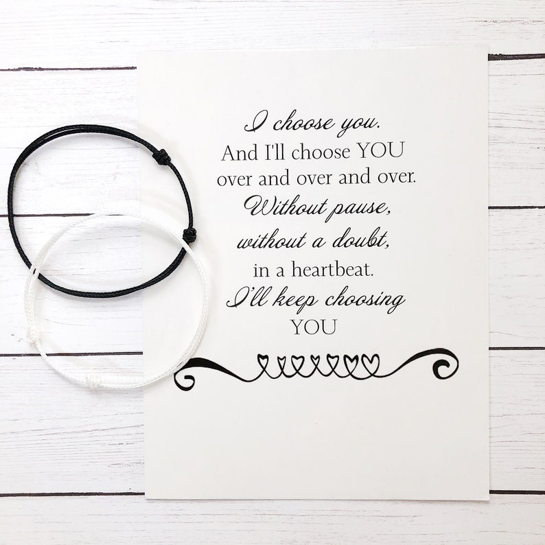 May include: A white card with black text that reads "I choose you. And I'll choose YOU over and over and over. Without pause, without a doubt, in a heartbeat. I'll keep choosing YOU" with a decorative heart design at the bottom. Two friendship bracelets, one black and one white, are on the left side of the card.