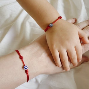 May include: Two red string bracelets with blue evil eye charms. The bracelets are worn on the wrists of two people, one adult and one child.