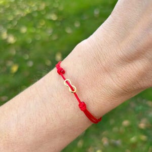 May include: Red string bracelet with a gold infinity symbol charm. The bracelet is tied with knots and is worn on a wrist. The background is a blurred green.