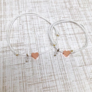 May include: Two white cord bracelets with rose gold heart charms. The bracelets are tied with a knot and have a small bead at the end.