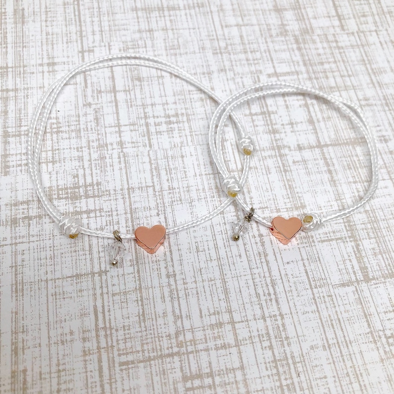 May include: Two white cord bracelets with rose gold heart charms. The bracelets are tied with a knot and have a small bead at the end.