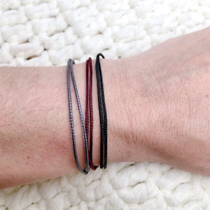 May include: A set of three bracelets in grey, burgundy, and black. The bracelets are made of thin cord and are tied together in a knot.