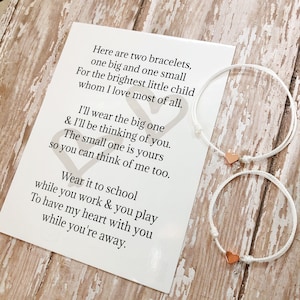 May include: Two white string bracelets with rose gold heart charms. The bracelets are accompanied by a card with a poem about wearing one bracelet and thinking of the person who wears the other.