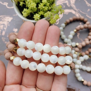 May include: Three beaded bracelets. Two bracelets feature white, round beads with gold-colored accents. The third bracelet has brown wooden beads. The bracelets are displayed in a hand.