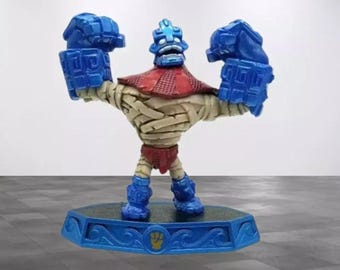 May include: A blue, red, and tan action figure of a muscular character with large blue fists and feet. The figure has a blue head and a red collar. It stands on a blue base with a gold emblem.