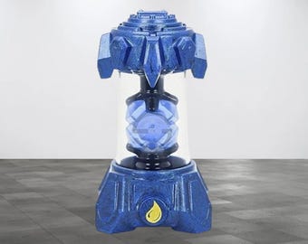 May include: A blue and silver toy with a clear plastic container holding a blue and silver sphere. The toy has a yellow oil drop symbol on the bottom.