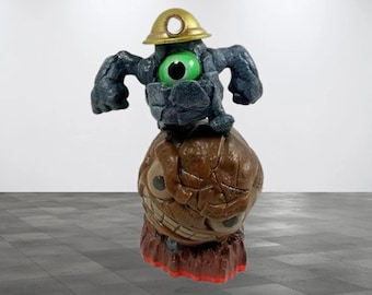 Può includere: Una statuetta stravagante di un personaggio dei cartoni animati. La figura ha un corpo grigio scuro, un grande occhio verde e un elmetto dorato. Si erge su una base marrone, strutturata, con un volto sorridente e dettagli rossi.