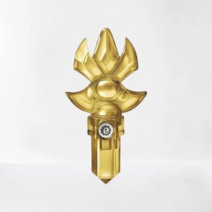 May include: A gold-colored, stylized key-shaped object with a geometric design. The top features a crown-like shape, and the base has a crystal-like point. A small circular emblem with a gear is visible near the base.