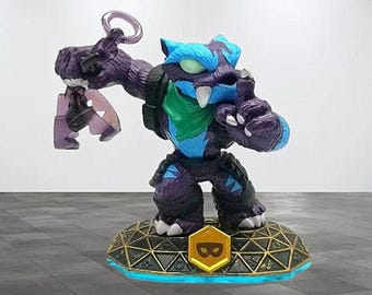 May include: A purple and blue action figure of a creature with a green scarf, standing on a gold and blue base. The figure has a claw-like hand and a ring on its other hand. The base has a gold emblem with a black symbol.