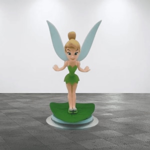 May include: A green and blue figurine of Tinkerbell, a fairy with wings, standing on a green leaf. The figurine is on a white base.