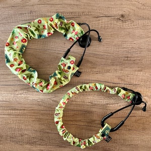 May include: Two stethoscope covers made of green fabric with a floral pattern. The covers are designed to fit around the stethoscope tubing and have a button closure.