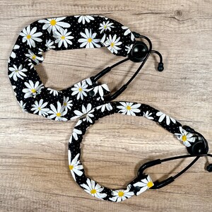 May include: Two black stethoscopes with daisy-patterned covers. The covers feature white daisies with yellow centers on a black background with white polka dots. The stethoscopes are on a light brown wooden surface.