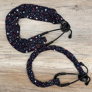 May include: Two black stethoscope covers featuring a repeating pattern of small white, red, and blue stars. Each fabric cover is gathered with elastic and includes a small black snap button closure to secure it around the stethoscope tubing.